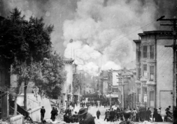 A black-and-white image showing people on a street with debris, buildings in disrepair, and smoke billowing in the background.