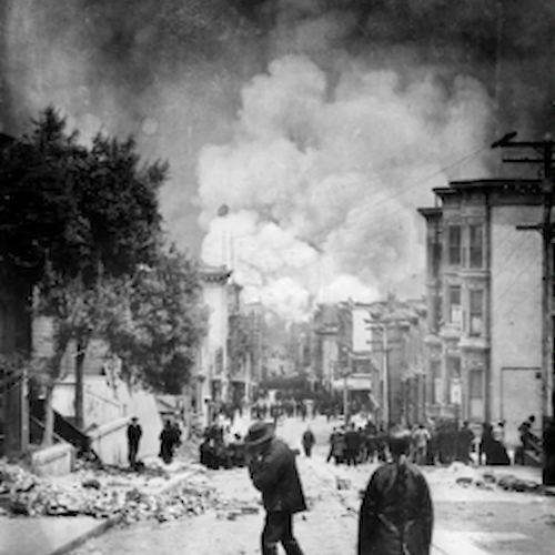 A black-and-white image showing people on a street with debris, buildings in disrepair, and smoke billowing in the background.