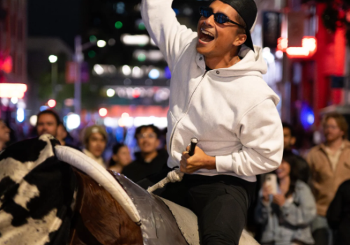 A person rides a mechanical bull, surrounded by a crowd on a city street at night, raising an arm in excitement while wearing sunglasses.