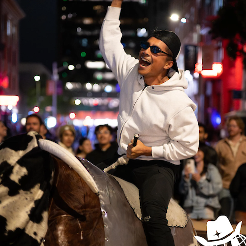 A person rides a mechanical bull, surrounded by a crowd on a city street at night, raising an arm in excitement while wearing sunglasses.