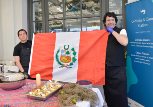 Two people hold a Peruvian flag by a table with food dishes and a sign saying "Cebiche & Causa Station" in the background.
