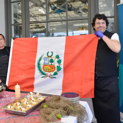 Two people hold a Peruvian flag by a table with food dishes and a sign saying "Cebiche & Causa Station" in the background.