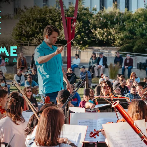 A conductor leads the Awesome Orchestra in an outdoor concert at Thrive City, surrounded by a large audience in a lively setting.