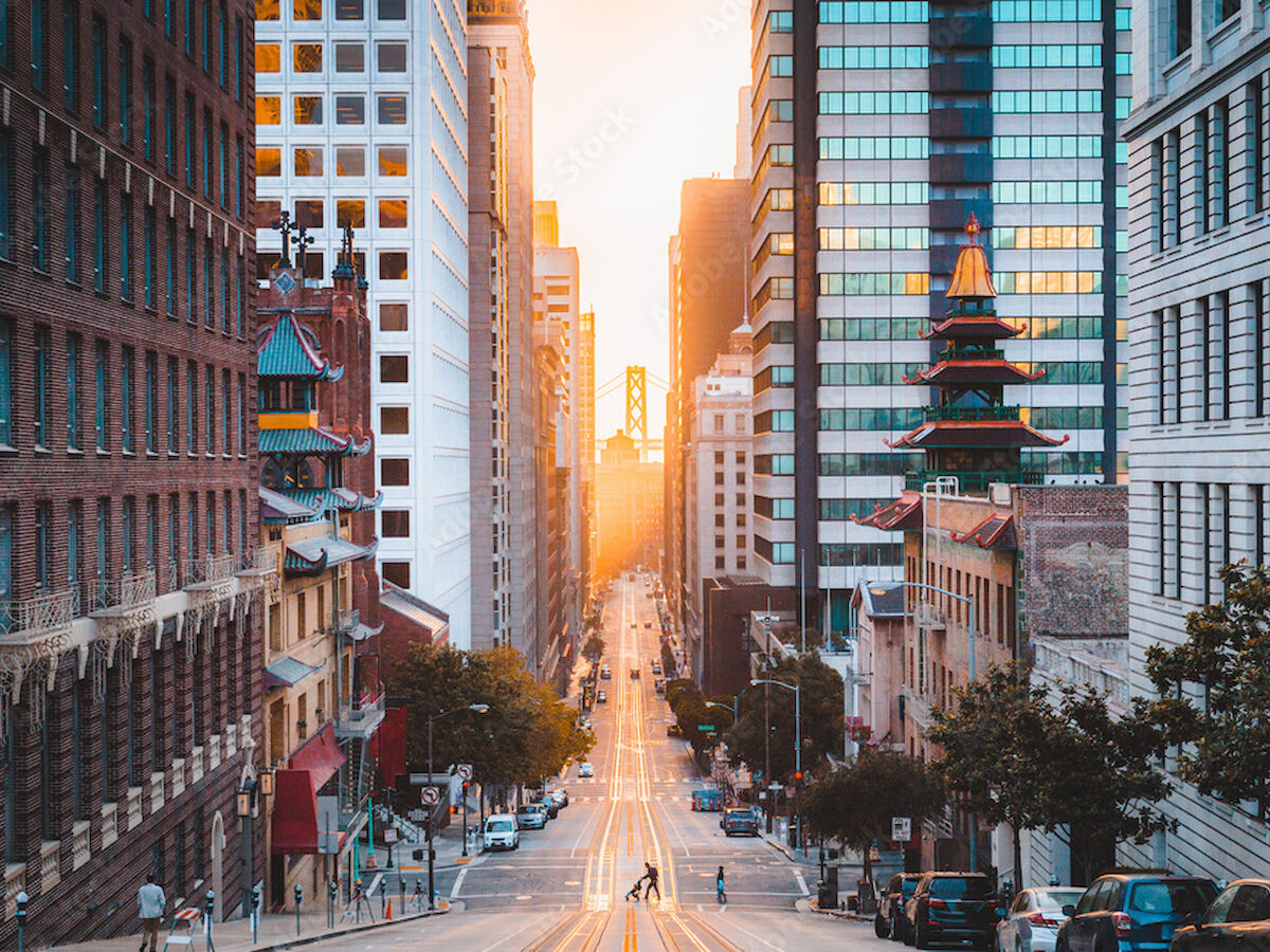 A city street flanked by tall buildings leading to the horizon, with a warm sunset and a glimpse of the Golden Gate Bridge in the distance.