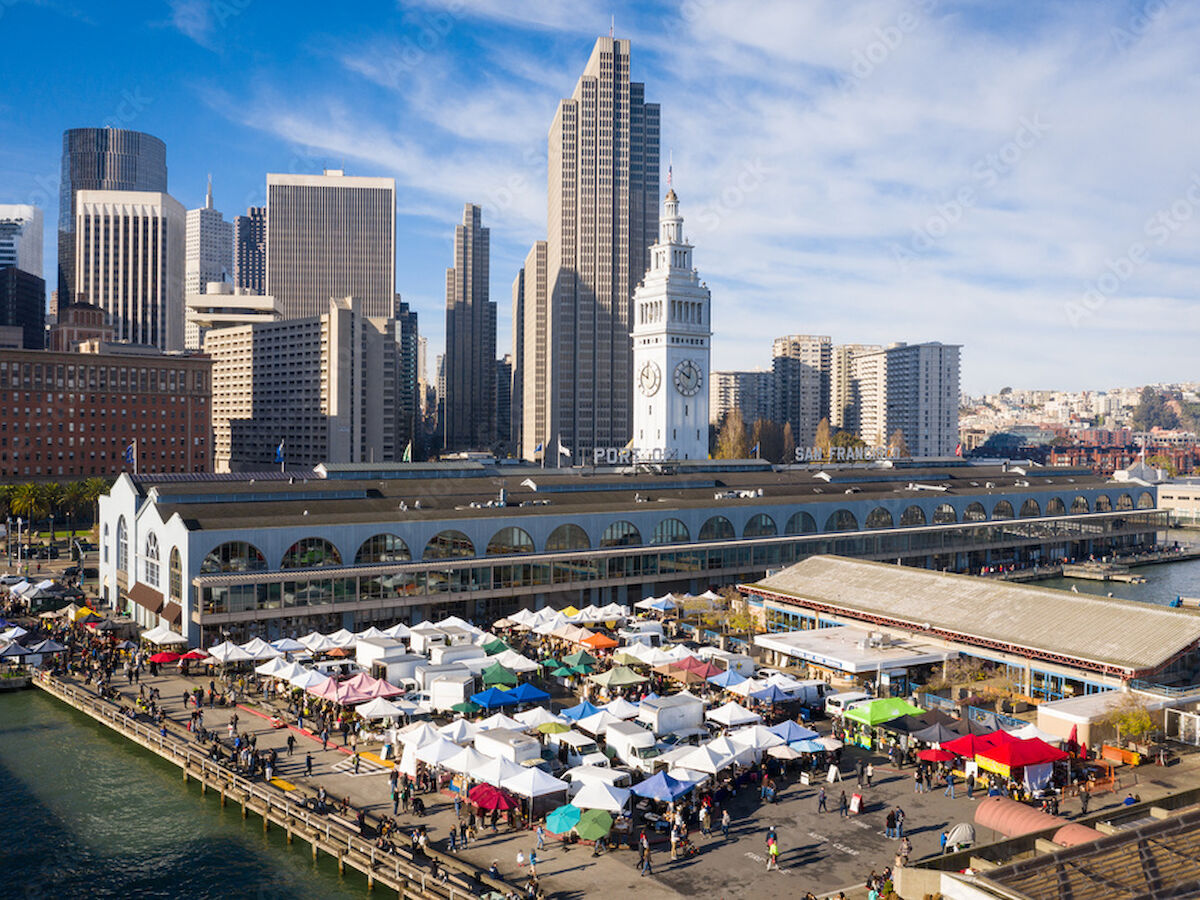 An outdoor market with white tents is set up near a waterfront, against a backdrop of tall city buildings and a clock tower.