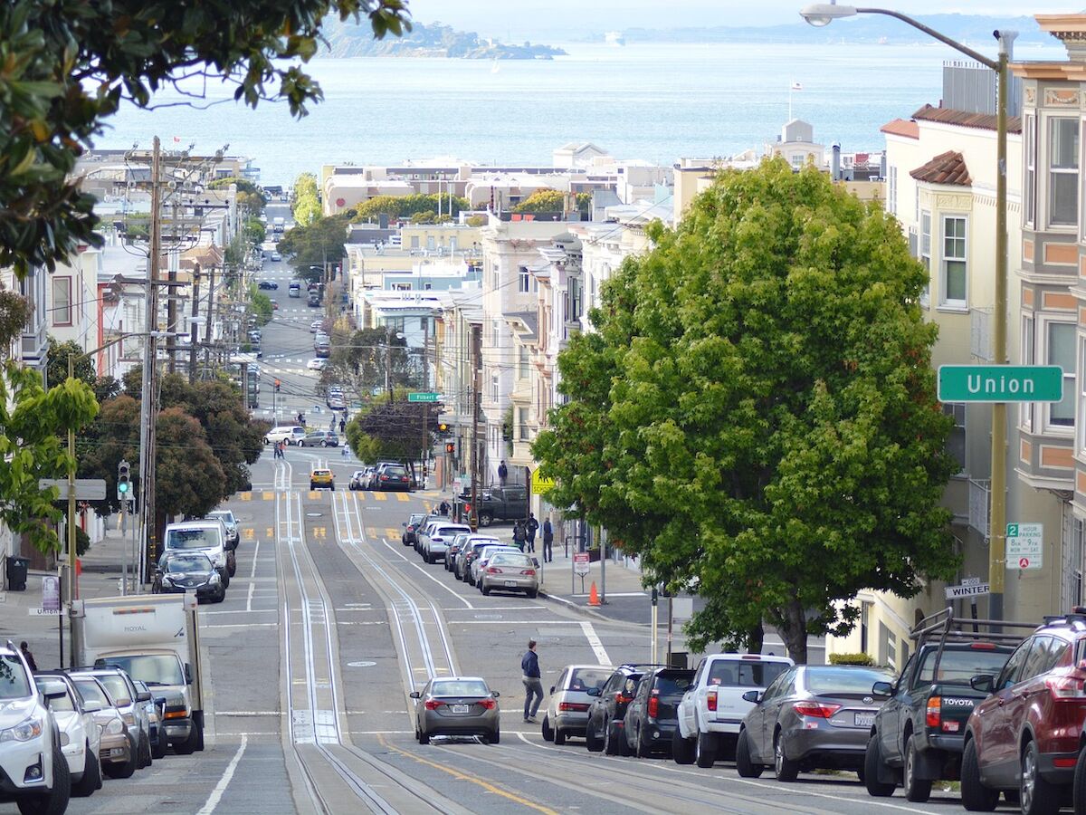 A sloped street with parked cars on both sides, a "Union" sign, people walking, and a view of the bay in the background.