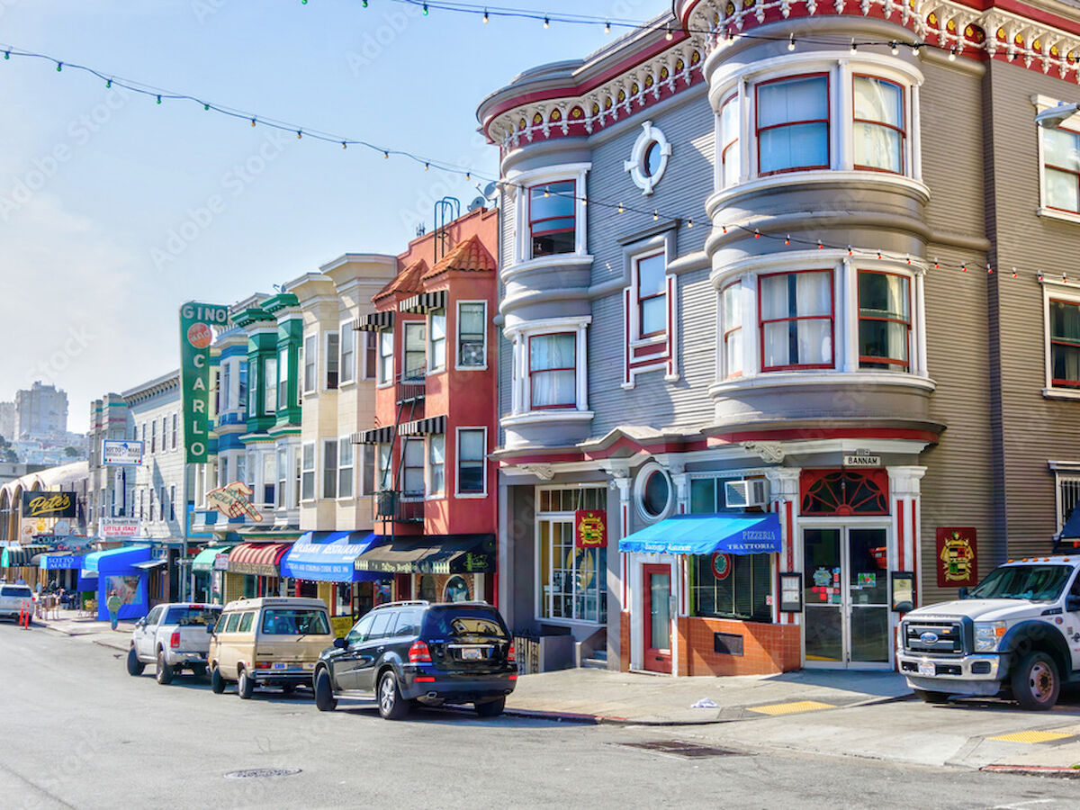 A street scene with colorful buildings, parked cars, and a sign for a restaurant. Some storefronts have awnings and decorative lights.