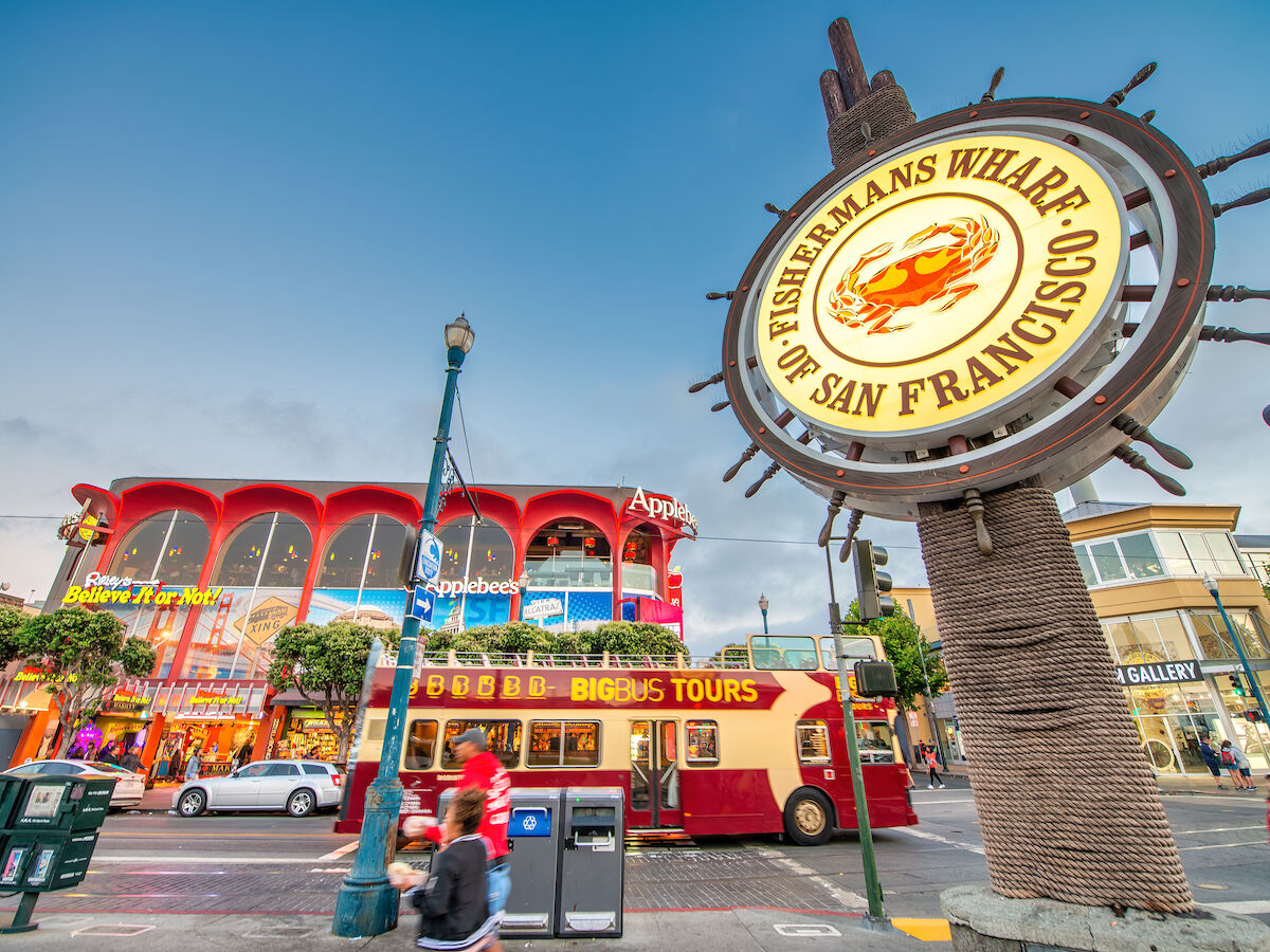 The image shows the iconic Fisherman's Wharf sign in San Francisco with a red double-decker bus and a colorful building in the background.