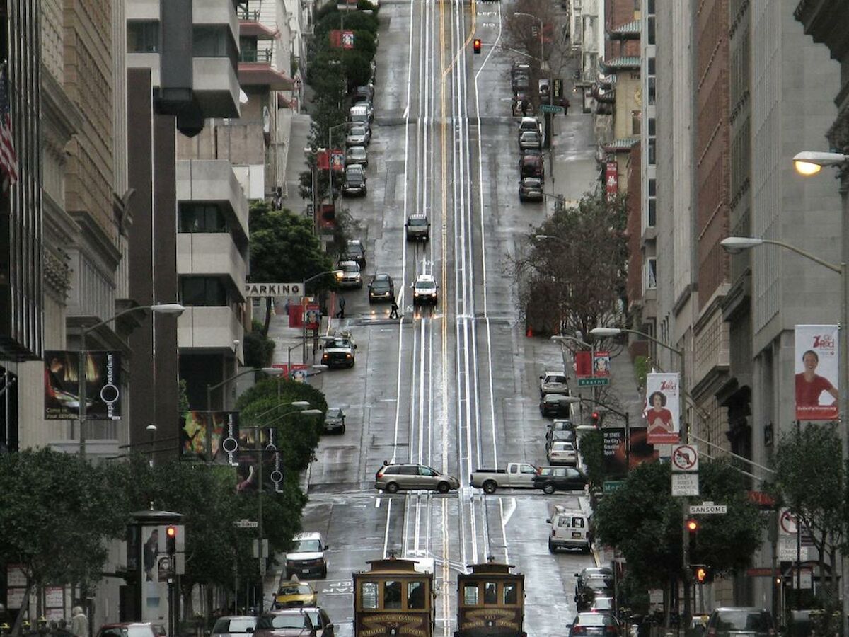 A steep, busy street lined with tall buildings, featuring cable cars and cars, likely in a hilly urban area.