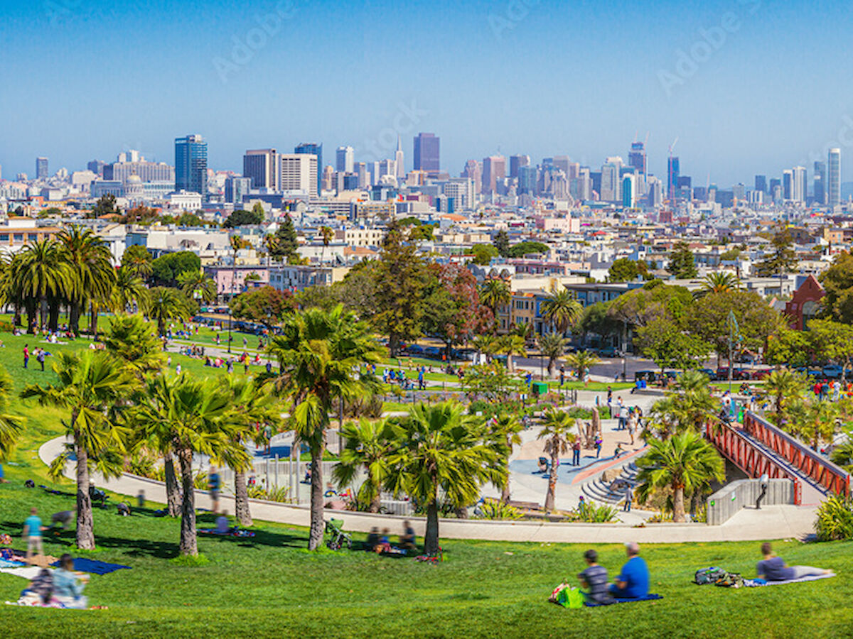 A park with people relaxing and a city skyline in the background, featuring palm trees and green spaces under a clear blue sky.