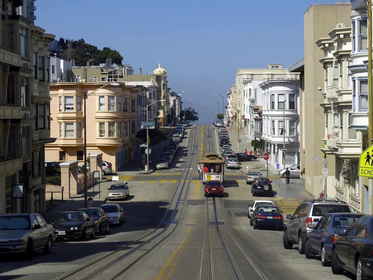 A street scene with a classic tram on hilly tracks, surrounded by rows of parked cars and city buildings under a clear blue sky.