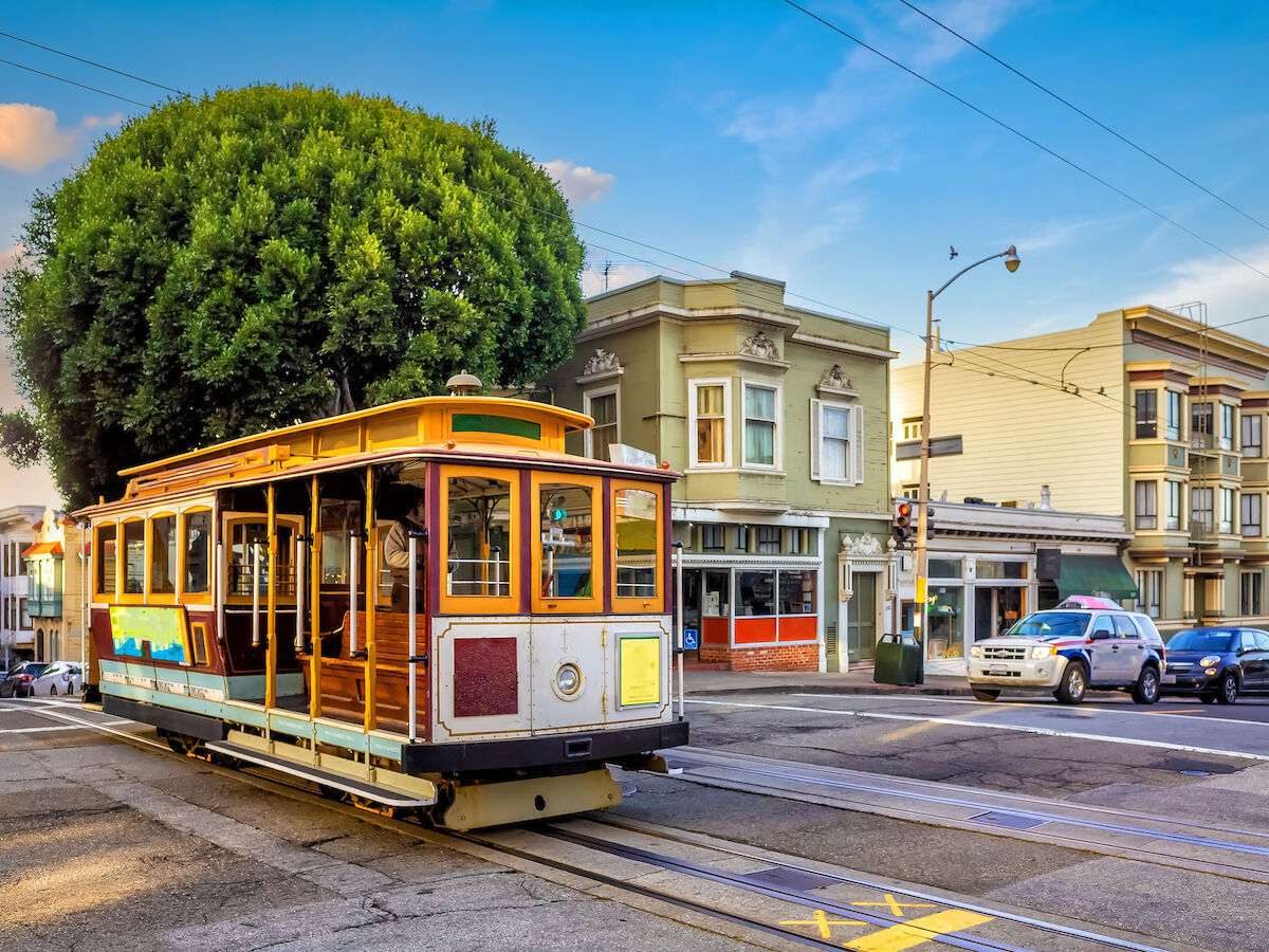 A vintage cable car on a city street with classic buildings and a large tree in the background, under a bright blue sky.