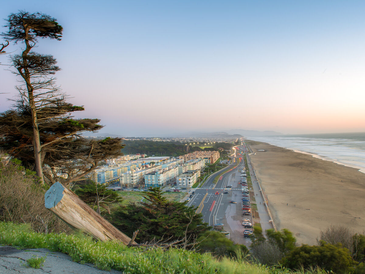 A coastal view with a road, beach, buildings, and trees at sunset. The scene has a peaceful, expansive horizon at dusk.