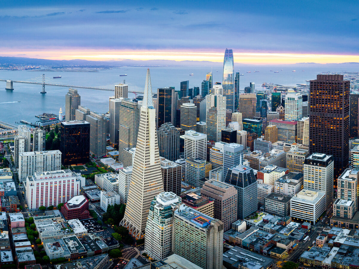 Aerial view of San Francisco skyline at sunset with tall buildings and the bay featuring the Bay Bridge in the background.
