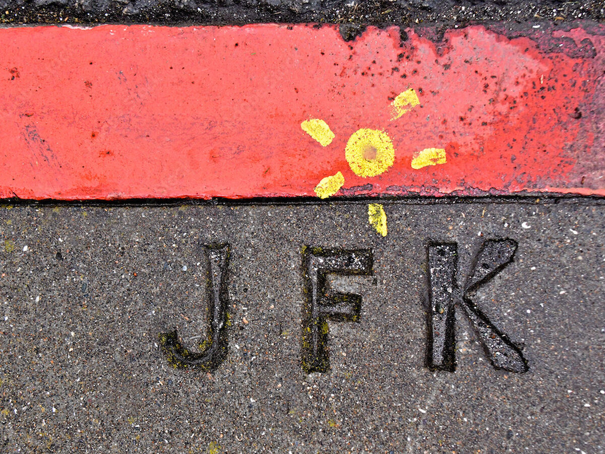 Concrete surface with "JFK" engraved and a red stripe, featuring a small yellow sun drawing.
