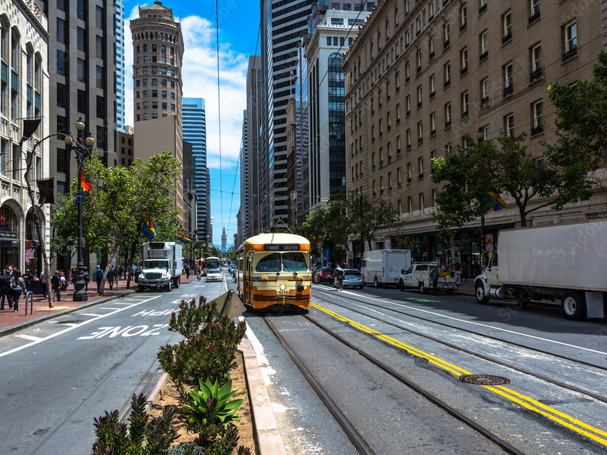 A street with a tram and tall buildings under a blue sky, with people walking and vehicles parked along the road.