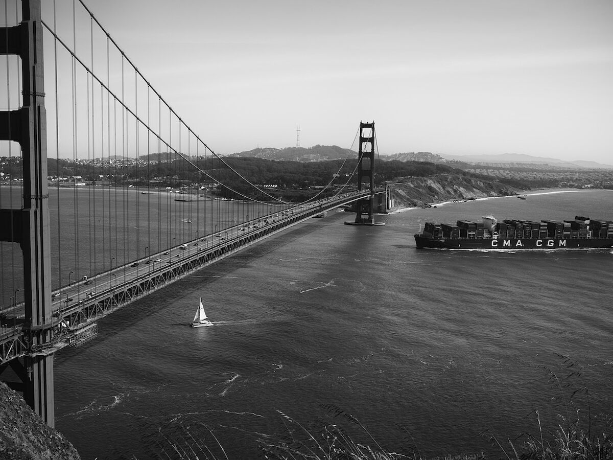 A black and white image of a suspension bridge with a ship passing underneath and a small sailboat on the water beside it.
