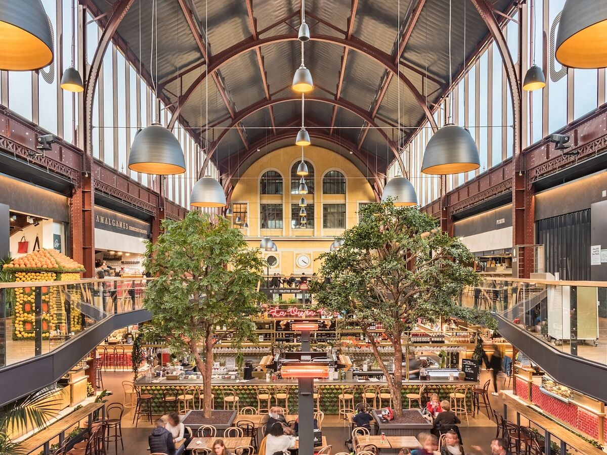 The image shows a large indoor market with seating, high ceilings, arches, plants, and people dining or shopping under large hanging lights.