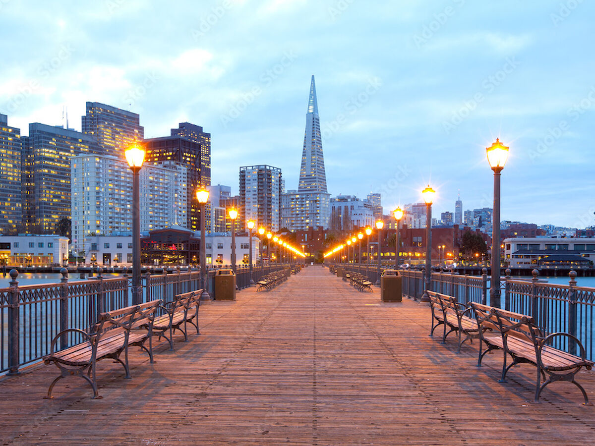 A wooden pier with benches leads to city skyscrapers, including a pointed building, illuminated by street lamps under a cloudy sky.