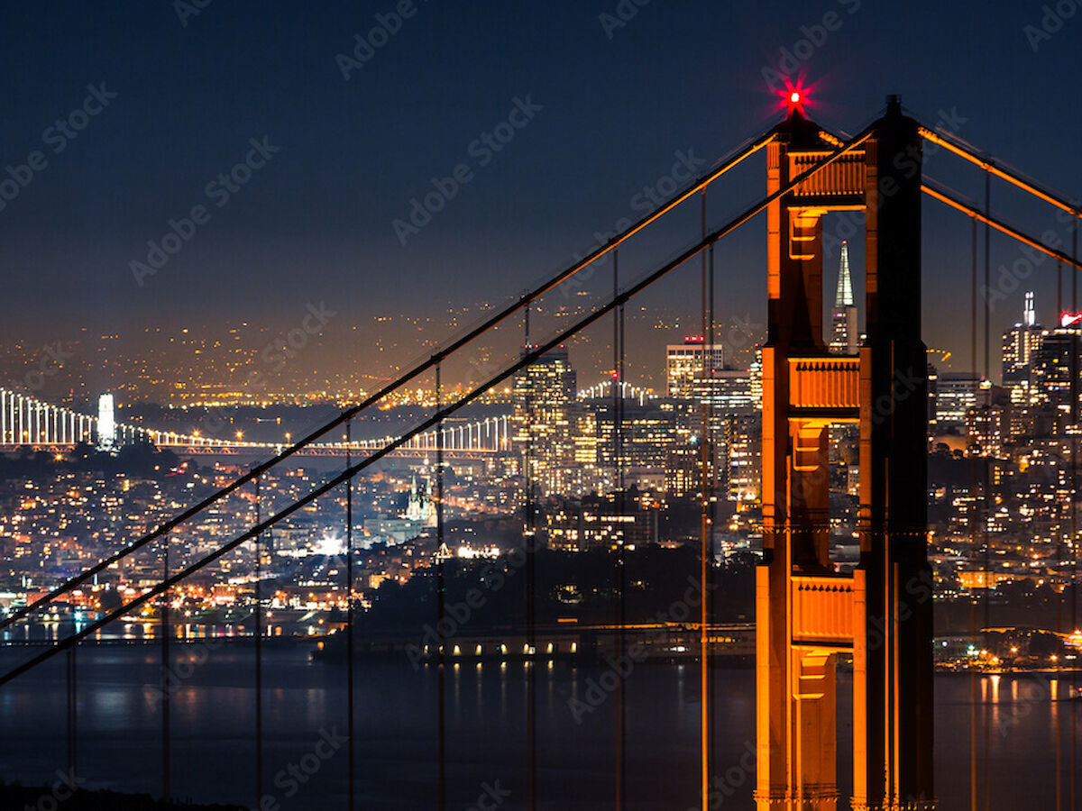 The image shows the Golden Gate Bridge illuminated at night with a cityscape in the background and reflections on the water.
