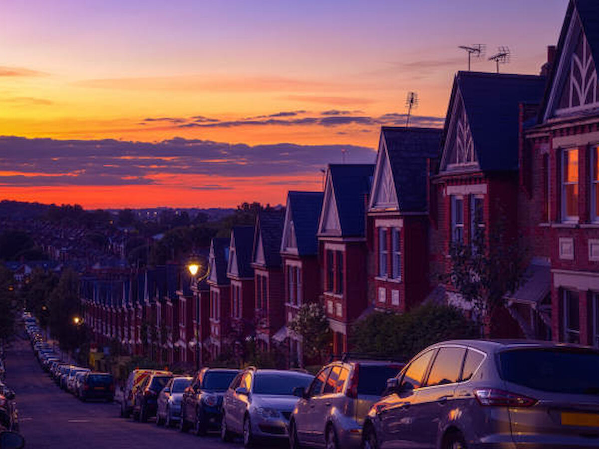 A row of red-brick houses lines a street at sunset, with cars parked along the curb and a vibrant sky in the background.