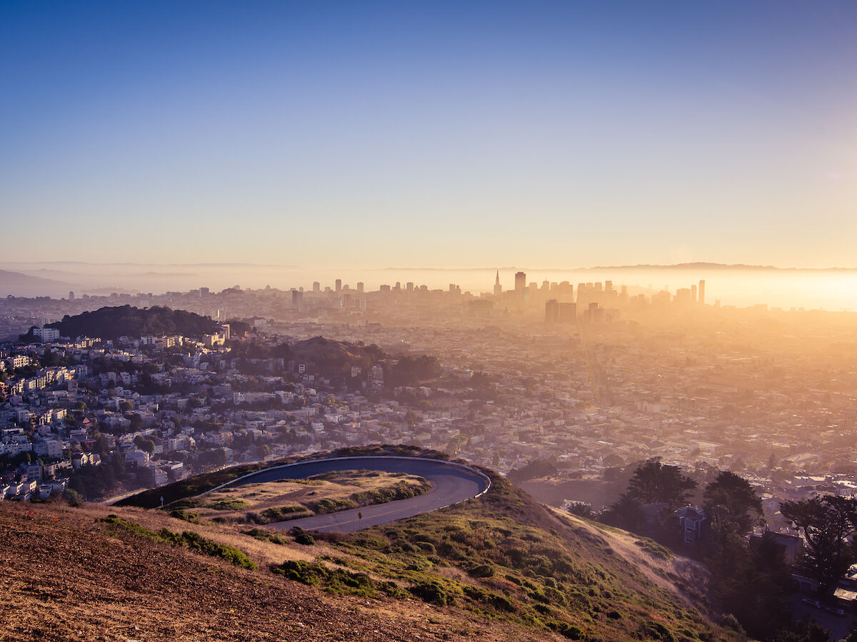 A cityscape at sunrise features a circular lookout on a hill and a skyline in the distance, enveloped in soft, golden light.