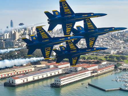 Four fighter jets fly in formation over a waterfront cityscape, producing trails of smoke against a clear blue sky.