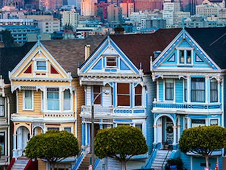 Colorful Victorian houses on a grassy hill with a city skyline in the background, featuring closely built, decorative architecture.