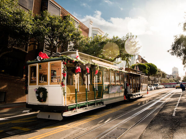 A decorated tram is traveling on a sunny street, with buildings and trees lining the background.