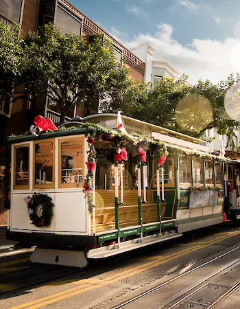A festive streetcar decorated with wreaths and ribbons travels down a city street lined with trees and buildings in daylight.