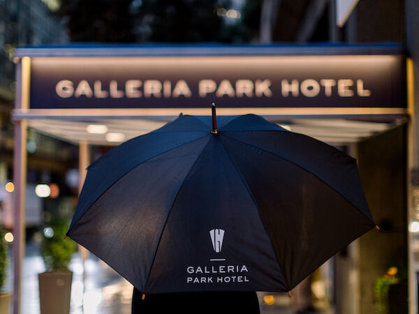 A large black umbrella with &ldquo;GALLERIA PARK HOTEL&rdquo; logo in front of the entrance, partially obscuring a sign that reads &ldquo;GALLERIA PARK HOTEL.&rdquo;