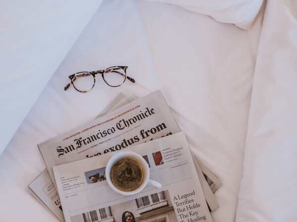 A pair of glasses rests on a newspaper titled &ldquo;San Francisco Chronicle&rdquo; with a coffee cup on top, all on white bedding.