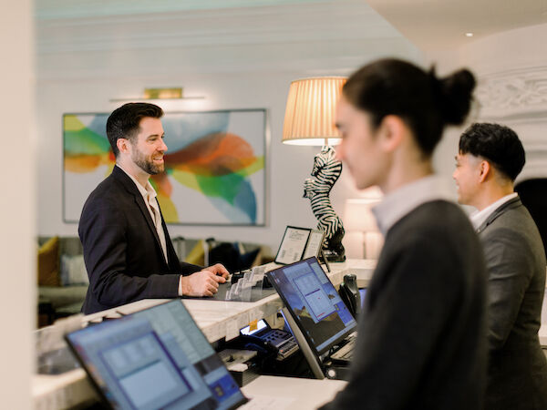 Two people at a counter assist a man in a suit, with computers and a colorful artwork in the background.