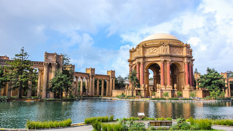 A large domed building with columns sits by a lake under a blue sky with clouds.