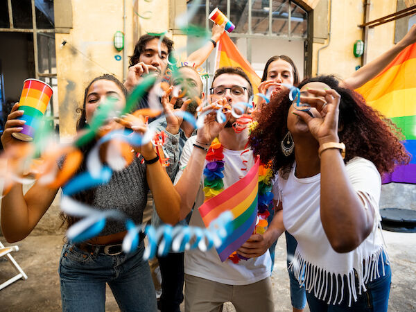 A group of diverse friends celebrate Pride with rainbow flags and banners, smiling and cheering in a lively outdoor setting in San Francisco