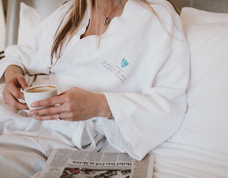 A person in a white robe sits in bed, holding a cup of coffee, with a newspaper on the lap.