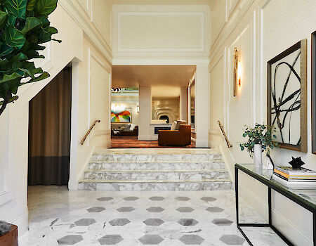 A bright, elegant lobby with a marble floor, steps to a seating area, modern art on the walls, a console table, and greenery.