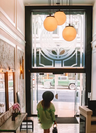 A person in a lime green coat walks toward a glass door in a stylish, bright lobby with hanging globe lights and marble walls.