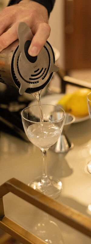 A hand pours a drink from a bottle into a cocktail glass on a bar, with other glasses and a lemon in the background.