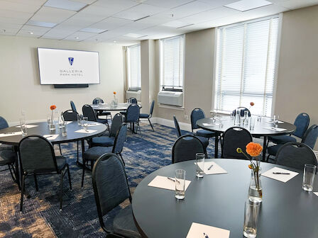 Hotel conference room with set round tables, blue chairs, orange flowers, big windows, and a wall TV reading &ldquo;Galleria Park Hotel&rdquo;.