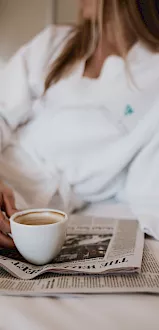 A person in a white shirt sipping from a coffee cup while reading an open newspaper at a table.