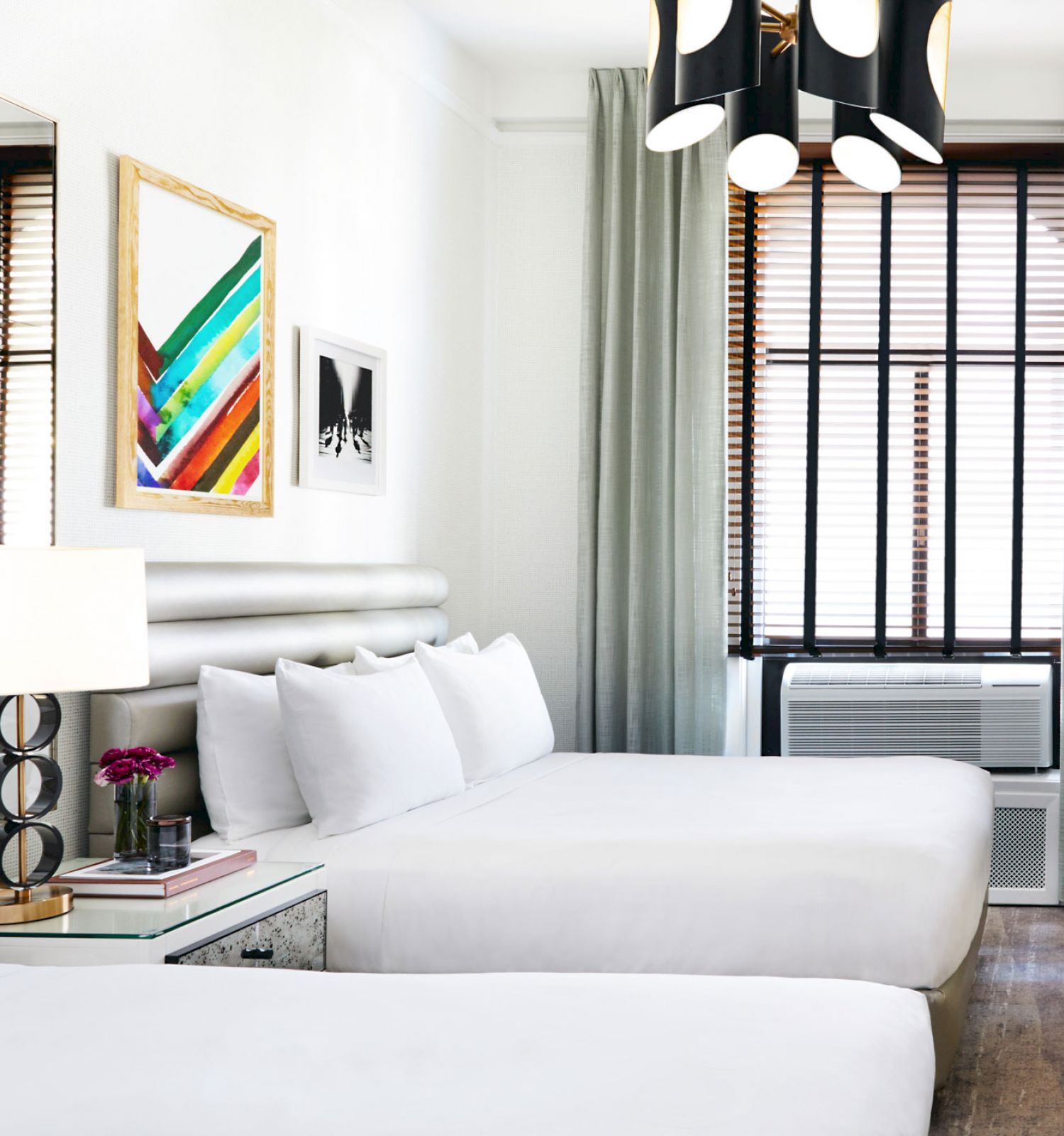 The image shows a modern hotel room with two beds, wall art, a lamp, curtains, a TV, and an air conditioning unit under the window with blinds.