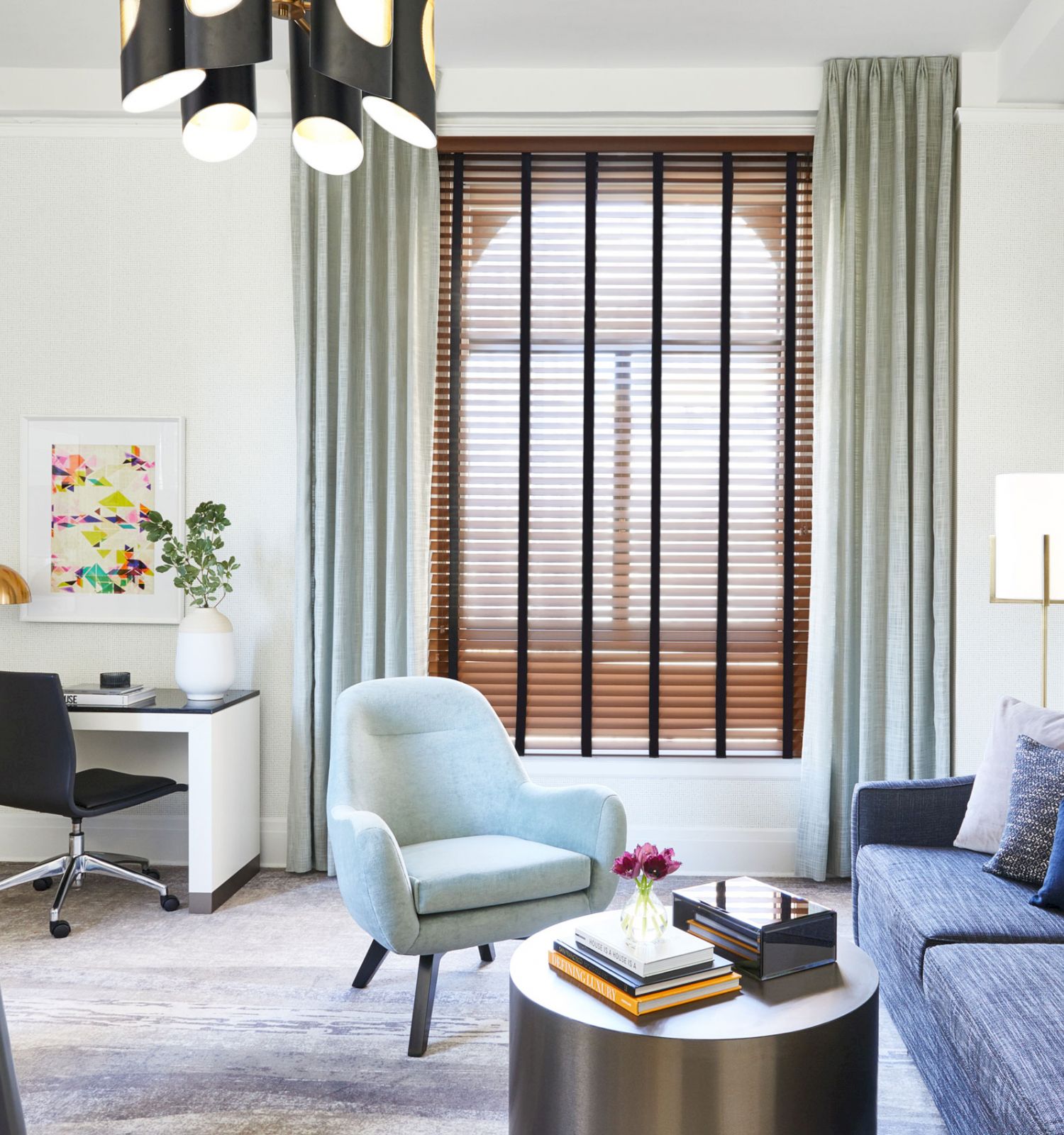 A modern living room with a desk, chair, sofa, armchair, coffee table, and wall art. The room is decorated in neutral tones with wooden blinds.