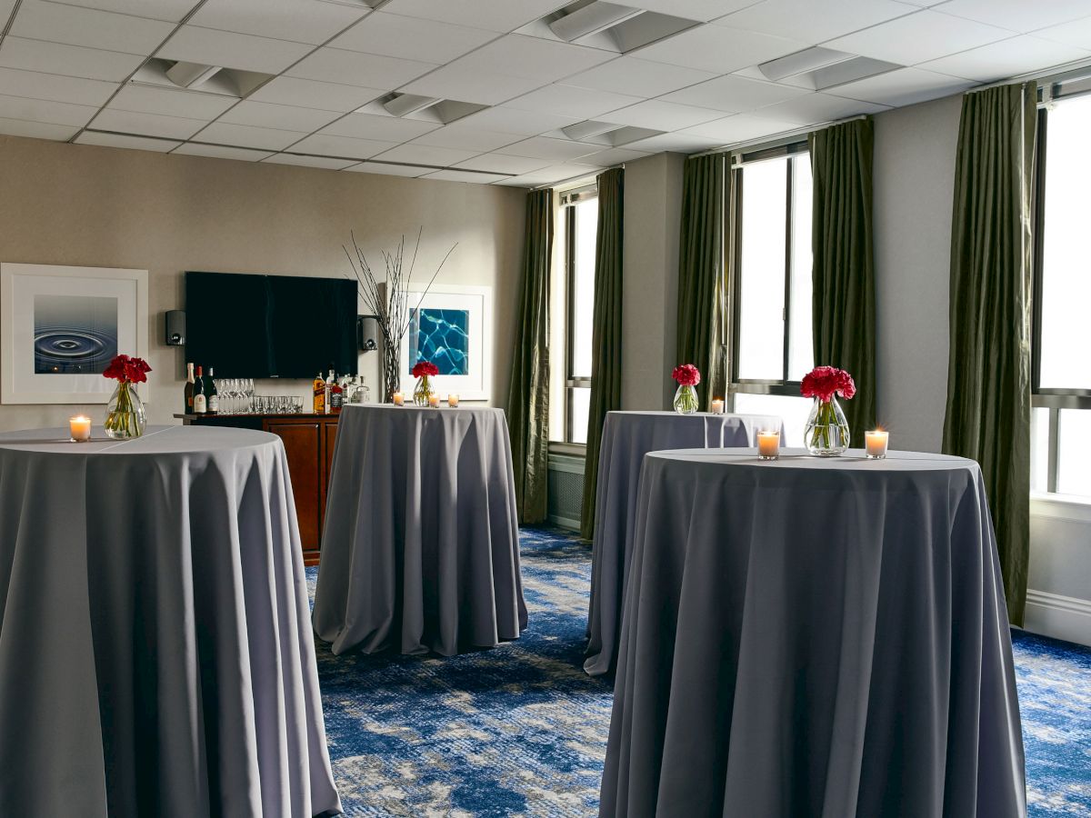 The image shows a room set up for an event with four tall tables draped in grey tablecloths, candles, and red flower arrangements on each table.