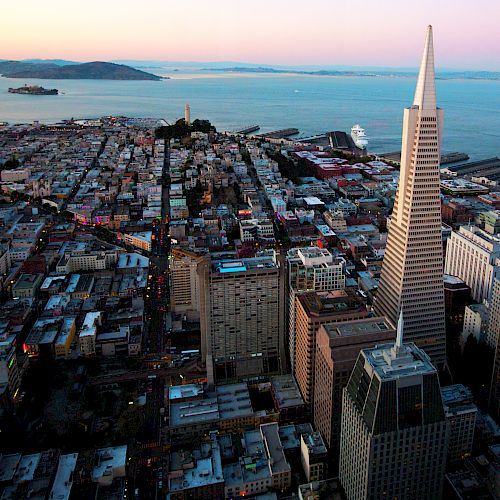 Aerial view of a coastal cityscape featuring a tall, pyramidal skyscraper and dense urban buildings with a body of water in the background.